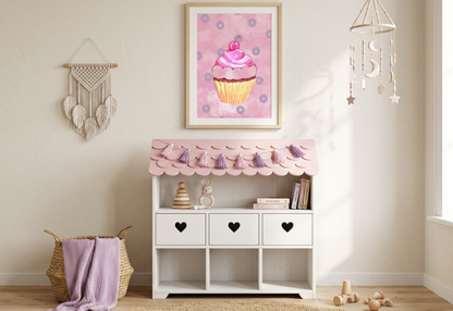 A photorealistic photograph of a serene, boho-themed nursery with cream-colored walls and a light wood floor covered by a round jute rug. In the center is a white wooden toy storage unit with a scalloped pink roof, featuring three drawers with heart-shaped cutouts and open shelving below. A tassel garland in shades of pink and purple is draped across the pink roof. Hanging directly above the unit is a light wood-framed vertical art print of a whimsical pink watercolor cupcake with purple donuts.
