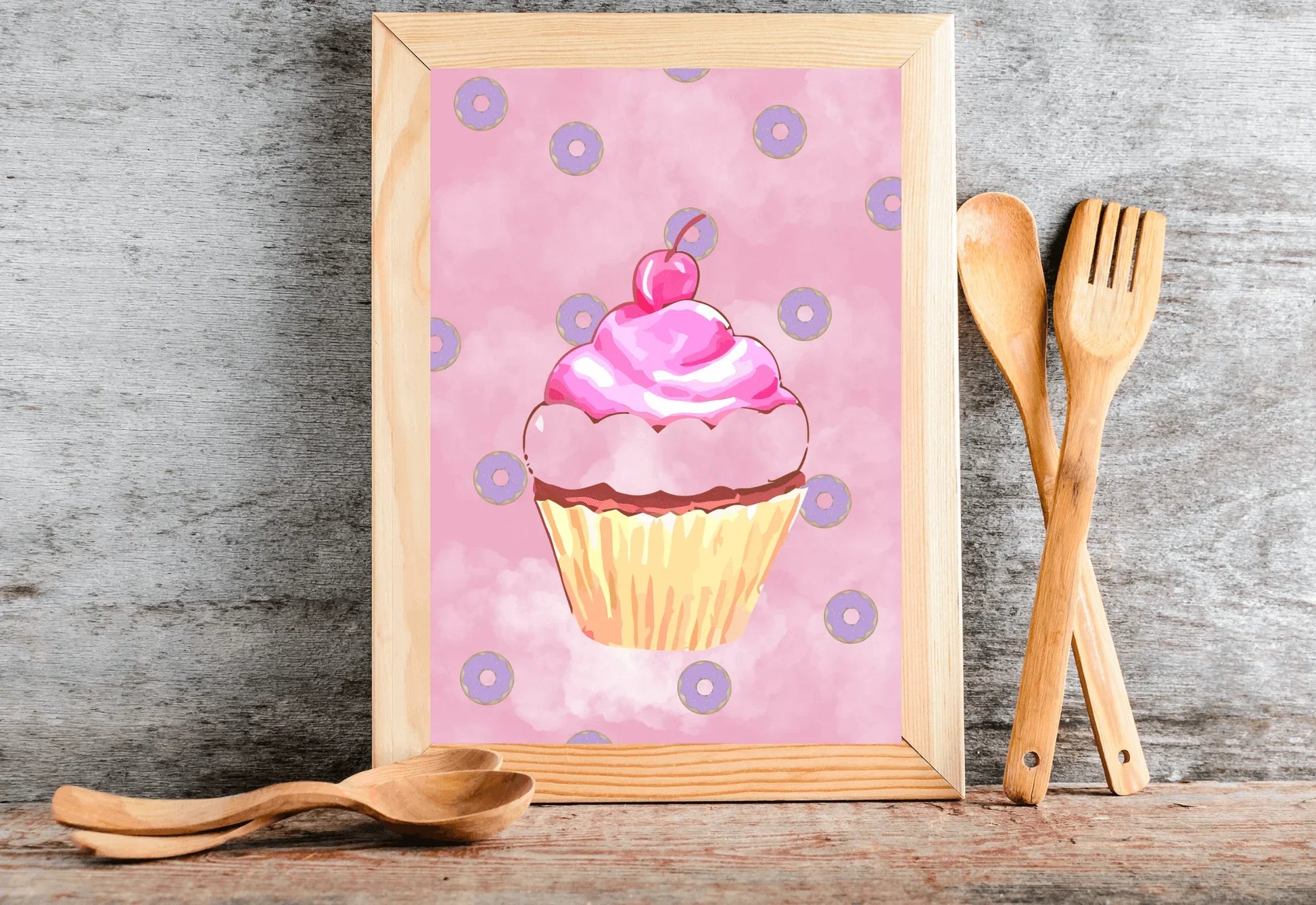A photorealistic photograph of a light wood-framed art print leaning against a textured grey stone wall. The print features a whimsical watercolor illustration of a pink cupcake with a cherry on top, surrounded by small purple donuts on a soft pink cloudy background. The frame sits on a rustic wooden surface, accompanied by two pairs of light-colored wooden spoons. One pair is laid horizontally in front of the frame, while the other pair is leaned vertically against the wall to the right. 