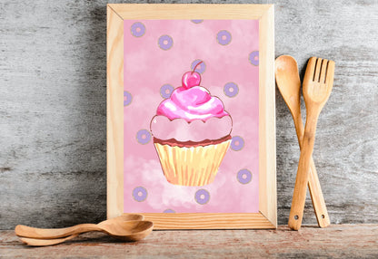 A photorealistic photograph of a light wood-framed art print leaning against a textured grey stone wall. The print features a whimsical watercolor illustration of a pink cupcake with a cherry on top, surrounded by small purple donuts on a soft pink cloudy background. The frame sits on a rustic wooden surface, accompanied by two pairs of light-colored wooden spoons. One pair is laid horizontally in front of the frame, while the other pair is leaned vertically against the wall to the right. 