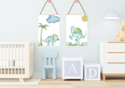 A set of two watercolor dinosaur art prints with wooden hangers on a light blue wall in a nursery with a crib and letter blocks.