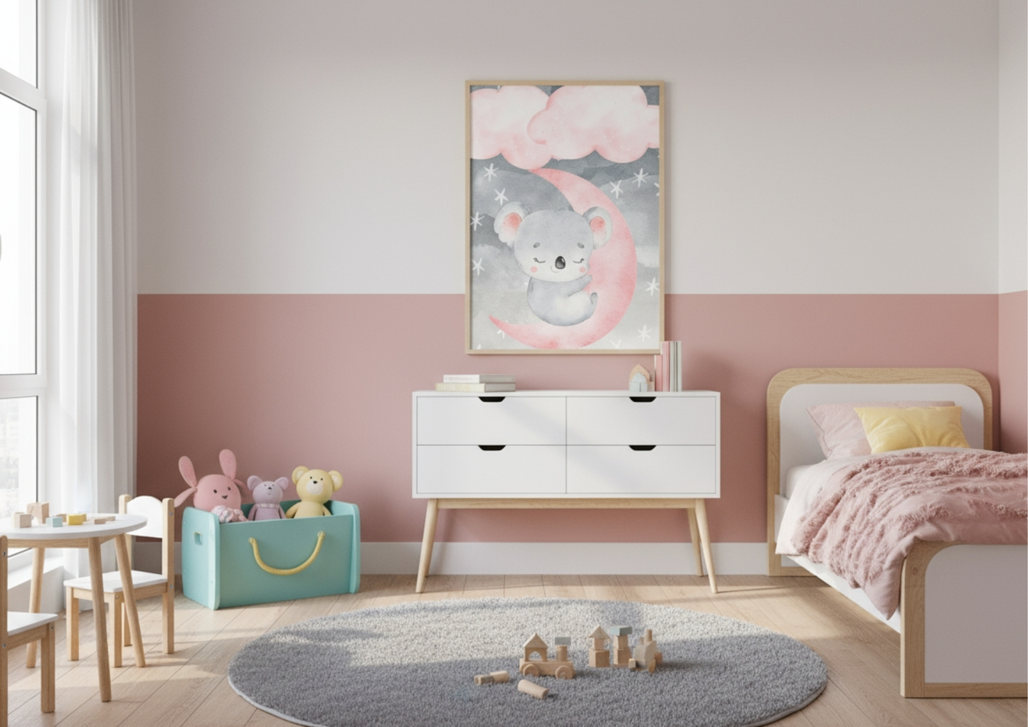 A bright, modern child's bedroom or nursery with walls painted in a two-tone design: white on the top half and a dusty rose pink on the bottom half. A framed watercolor print of a sweet, sleeping koala resting on a pink crescent moon, under clouds and stars, hangs centrally above a white mid-century modern dresser. To the right, a low wooden toddler bed is visible, dressed with a chunky pink knit blanket. The room features a small table and chairs set, a teal toy bin filled with stuffed animals, and a round