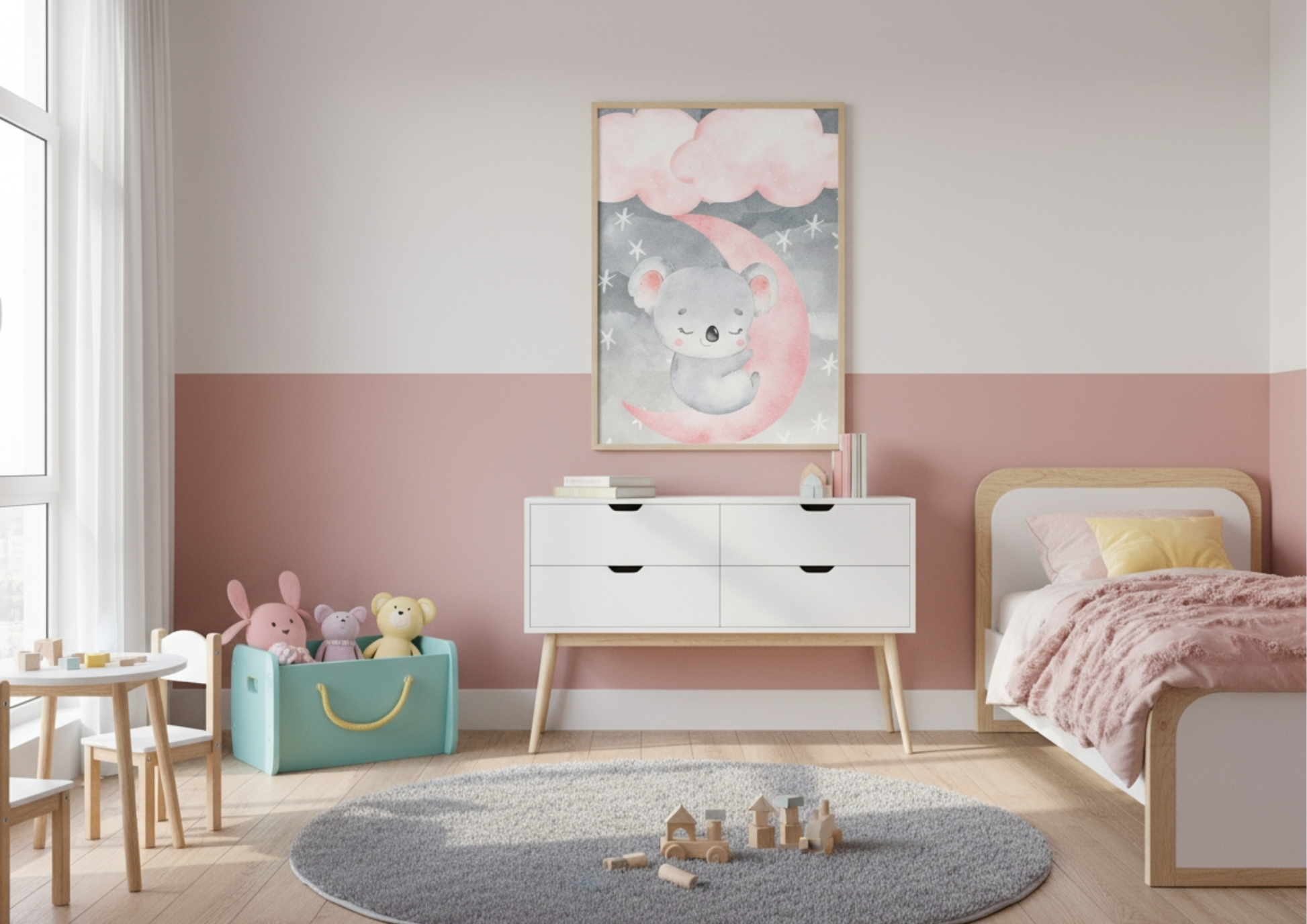A bright, modern child's bedroom or nursery with walls painted in a two-tone design: white on the top half and a dusty rose pink on the bottom half. A framed watercolor print of a sweet, sleeping koala resting on a pink crescent moon, under clouds and stars, hangs centrally above a white mid-century modern dresser. To the right, a low wooden toddler bed is visible, dressed with a chunky pink knit blanket. The room features a small table and chairs set, a teal toy bin filled with stuffed animals, and a round