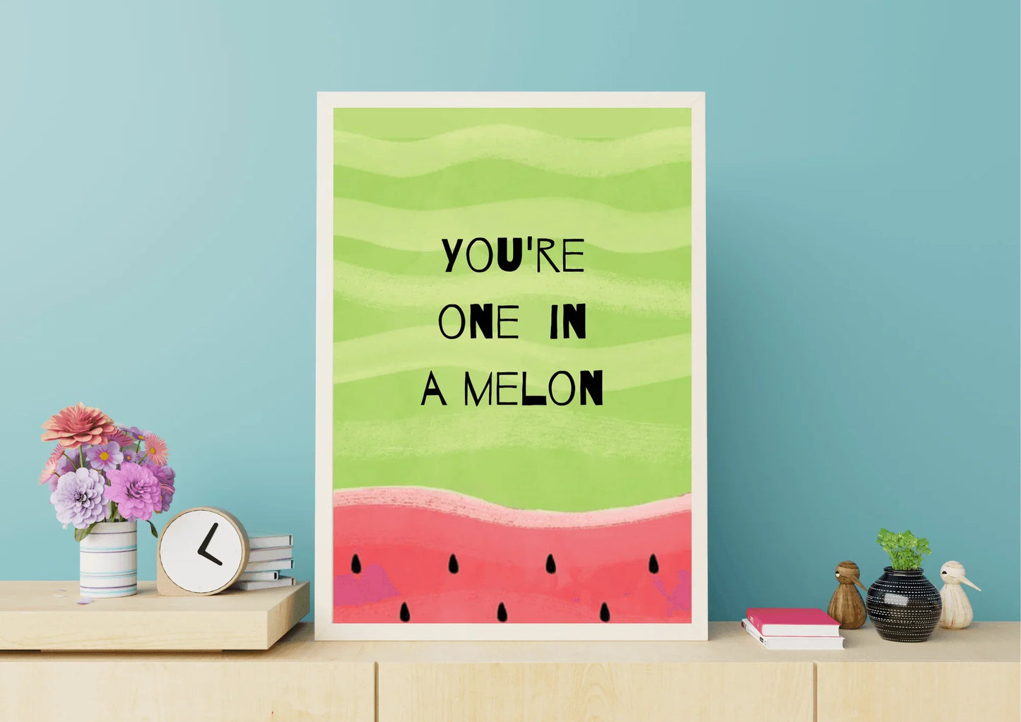 A framed, whimsical digital art print featuring a watermelon theme. The top two-thirds is a light green with wavy stripes, containing the black hand-drawn text "YOU'RE ONE IN A MELON". The bottom third is a textured pink/red color with black watermelon seeds. The print is displayed on a light wooden shelf against a teal wall, next to a small bouquet of flowers and a clock