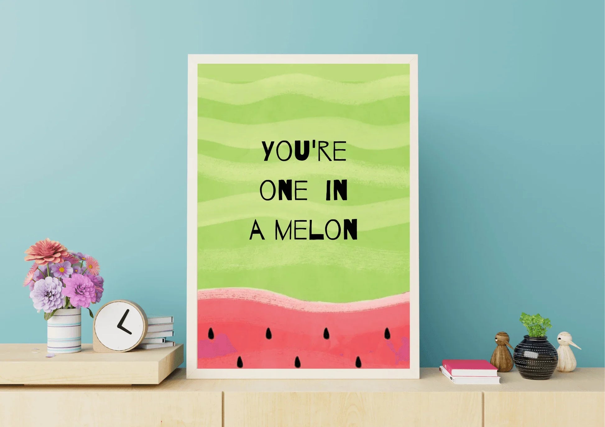 A framed, whimsical digital art print featuring a watermelon theme. The top two-thirds is a light green with wavy stripes, containing the black hand-drawn text "YOU'RE ONE IN A MELON". The bottom third is a textured pink/red color with black watermelon seeds. The print is displayed on a light wooden shelf against a teal wall, next to a small bouquet of flowers and a clock