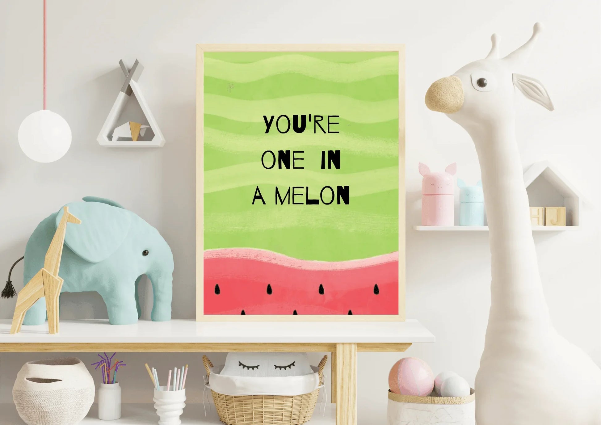 A framed "YOU'RE ONE IN A MELON" watermelon print, set on a white wooden table or dresser against a light wall in a child's room or nursery. The print is surrounded by toys and decor, including a large, soft white stuffed giraffe on the right, a turquoise blue stuffed elephant and a wooden toy on the left, and various small items on floating shelves above. A wicker basket with a smiling cloud design is visible below the table.