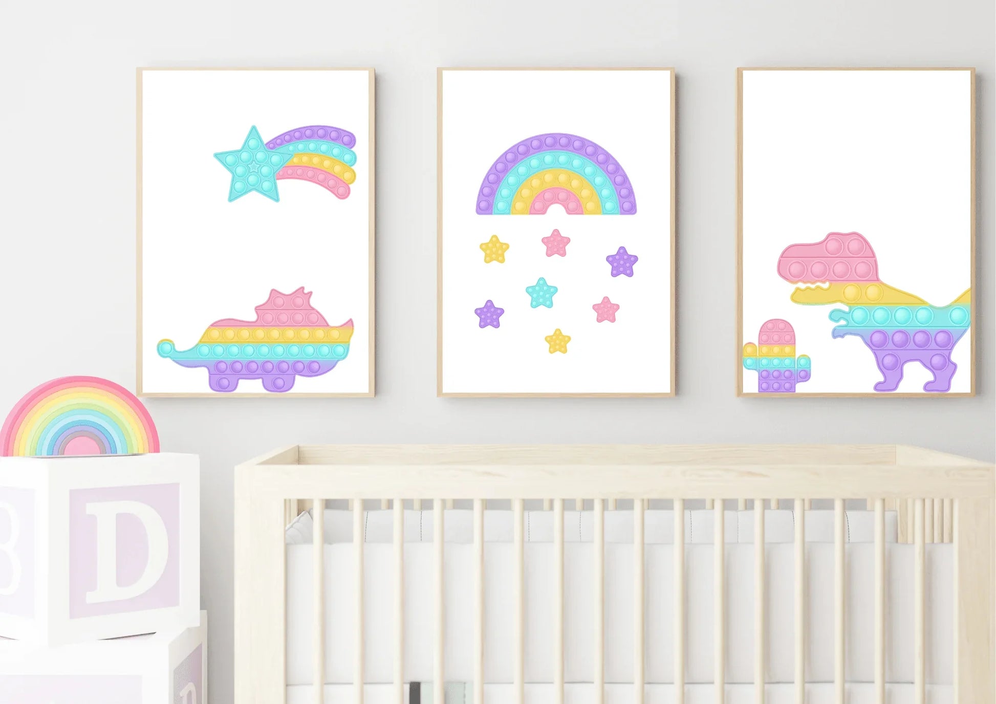 A three-piece nursery print set, featuring pop-it style dinosaurs, a rainbow, and a shooting star, hanging on a white wall above a wooden crib and a toy rainbow.